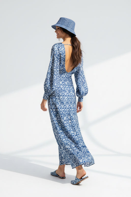 A woman wearing a blue and white patterned maxi dress with a deep open back, a matching blue bucket hat, and flat sandals, photographed against a bright white studio background.