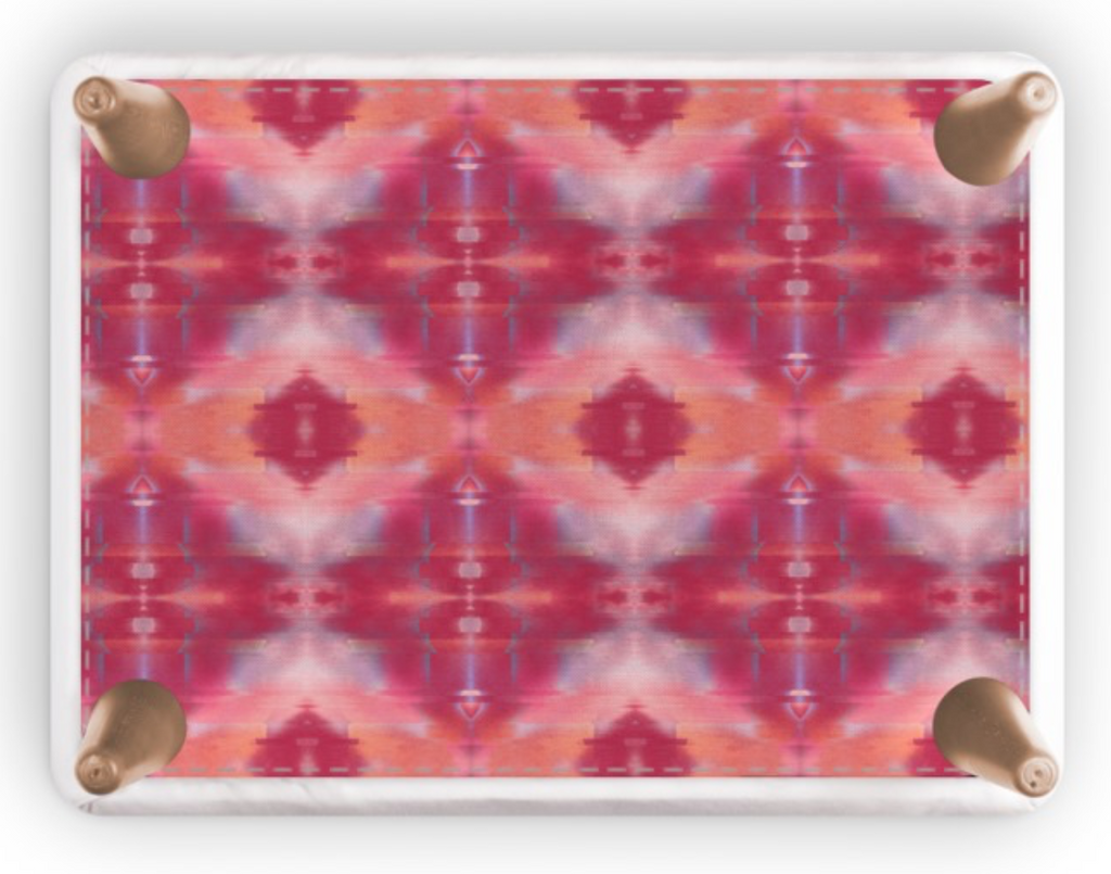 A rectangular footstool with rounded corners and four metallic rose gold feet, featuring a vibrant pink, red, and orange kaleidoscopic pattern on a white background.