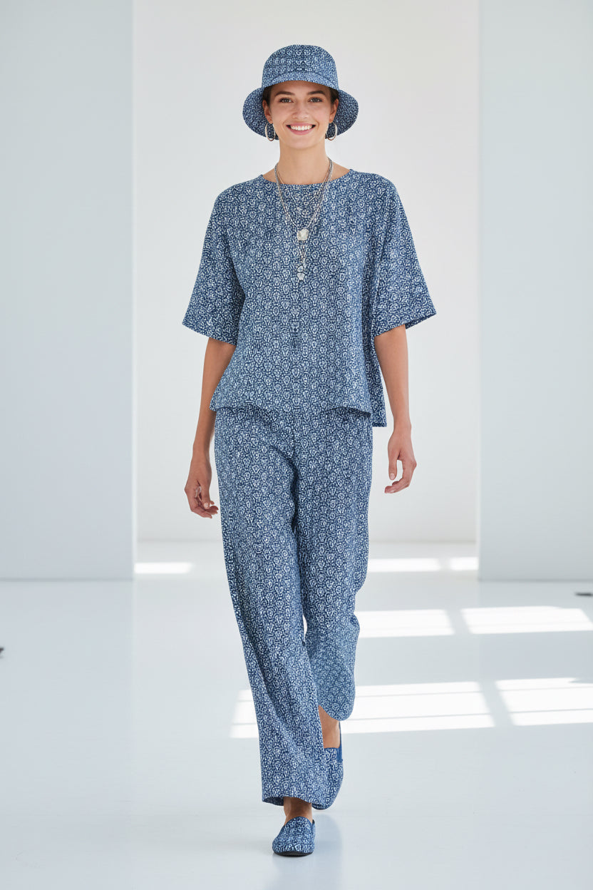 A woman modeling a matching blue and white patterned lounge set consisting of a short-sleeve top and wide-leg pants, along with a matching bucket hat and slip-on shoes, in a bright white studio setting.