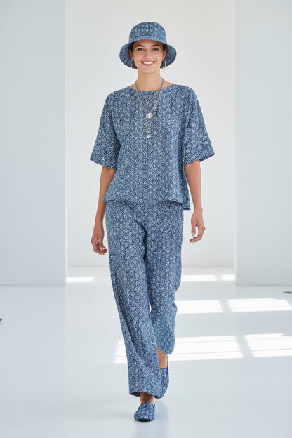 A woman modeling a matching blue and white patterned lounge set consisting of a short-sleeve top and wide-leg pants, along with a matching bucket hat and slip-on shoes, in a bright white studio setting.