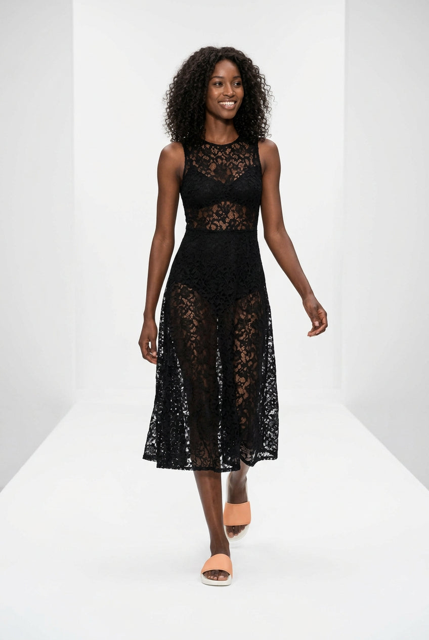 A woman walks down a white runway wearing a sleeveless black lace midi dress with a sheer lace bodice and skirt, paired with peach slide sandals.